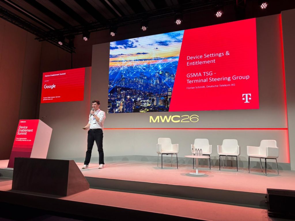 A man in a white shirt and jeans speaks on stage at MWC24. Behind him is a large screen showing “Device Settings & Entitlement,” with logos from Google and Deutsche Telekom. The stage has empty white chairs and a pink podium under bright lighting.