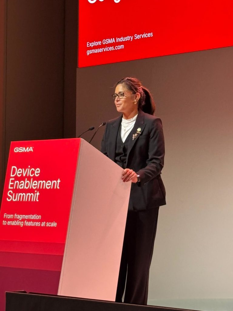 A woman in a black suit and white blouse stands at a podium labeled "Device Enablement Summit" against a red and gray backdrop. She wears glasses and a brooch, and smiles while speaking at the event. A large red screen with event information is in the background.