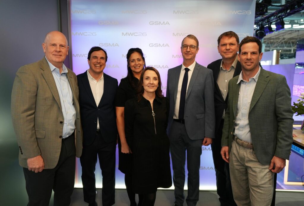 Seven people stand indoors in business attire, smiling at the camera. There are four men and three women. Behind them is a lighted backdrop with the text "GSMA" and "MWC23" repeated in pale blue and gold. The setting appears to be a professional event.
