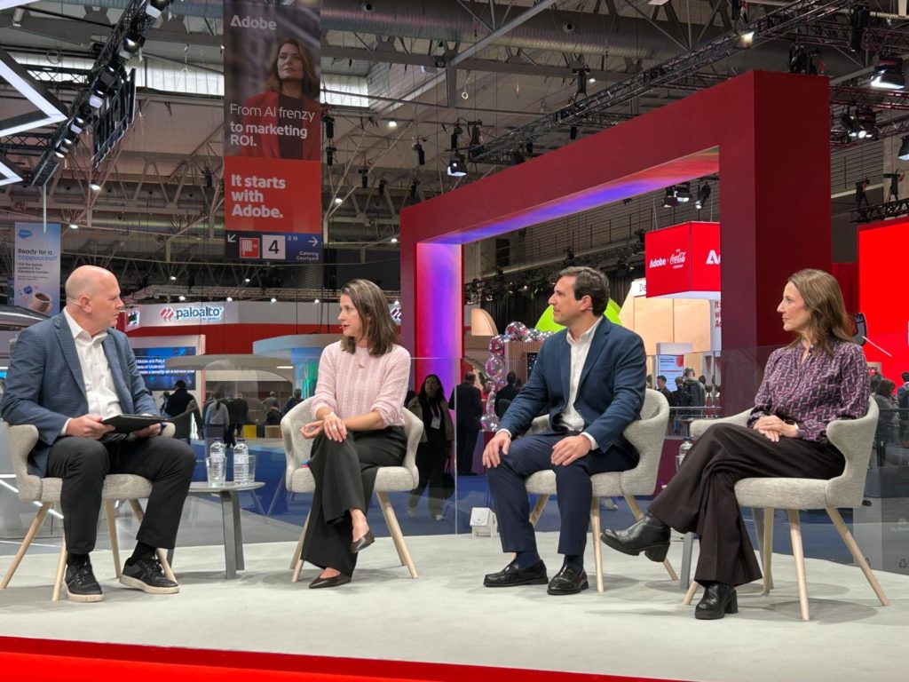 Four people sit on stage in modern chairs, engaged in a panel discussion. Behind them is a red frame structure and large digital screens, with various logos including Adobe. The setting is a professional event or conference, and the atmosphere is lively and focused.