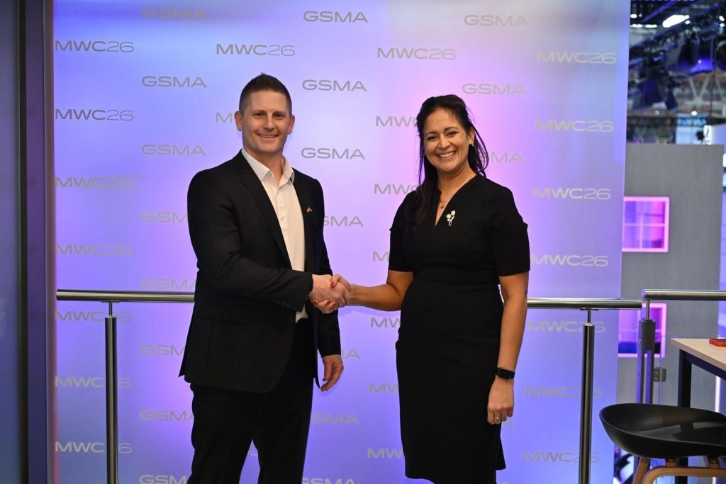 A man in a black suit and a woman in a black dress shake hands, smiling, in front of a purple backdrop with “GSMA” and “MWC26” logos. They stand indoors near rails, with modern event decor and lighting in the background.