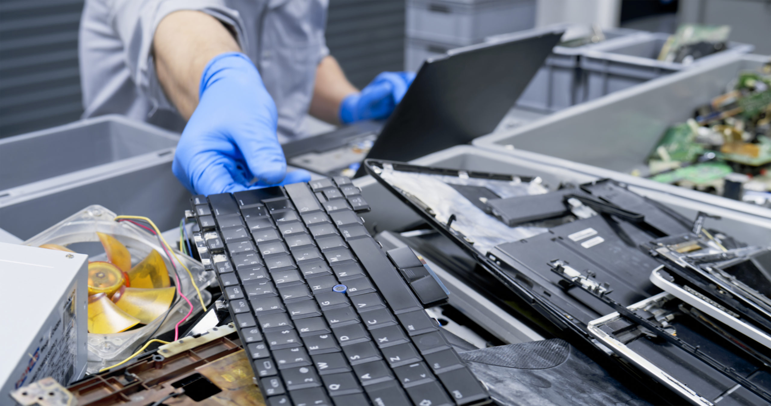 A person wearing blue gloves inspects and sorts electronic waste, including a detached keyboard and broken laptops, with a laptop in the background, highlighting an electronics recycling or repair facility.
