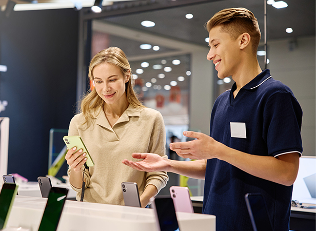 A woman smiles while holding a green smartphone in an electronics store. A young male employee in a navy shirt stands beside her, gesturing and talking. Several smartphones in different colors are displayed on the counter in front of them.