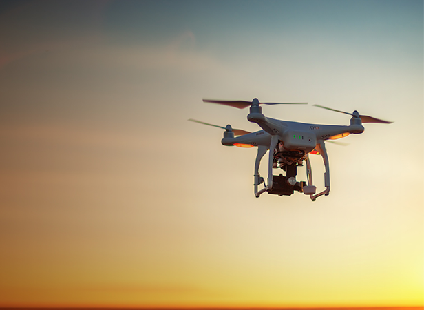 A white quadcopter drone with a camera hovers in midair against a colorful sunset sky, with orange and yellow hues fading into blue. The drone's propellers are spinning, and soft natural light highlights its body and camera attachment.