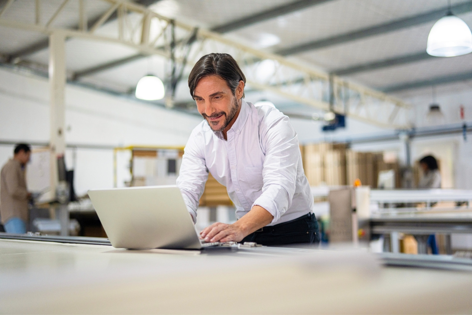 A man in a white shirt smiles while working on a laptop in a bright, spacious industrial setting. Machinery and other people blurred in the background suggest a busy, collaborative workplace. Overhead lights illuminate the scene.
