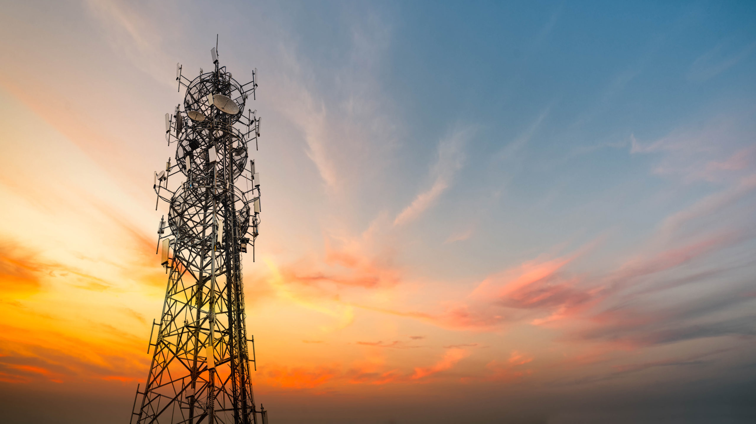 A tall telecommunications tower with numerous antennas stands against a dramatic sunset sky, featuring orange, yellow, and blue tones with wispy clouds. The silhouette of the metal structure contrasts with the colorful background.