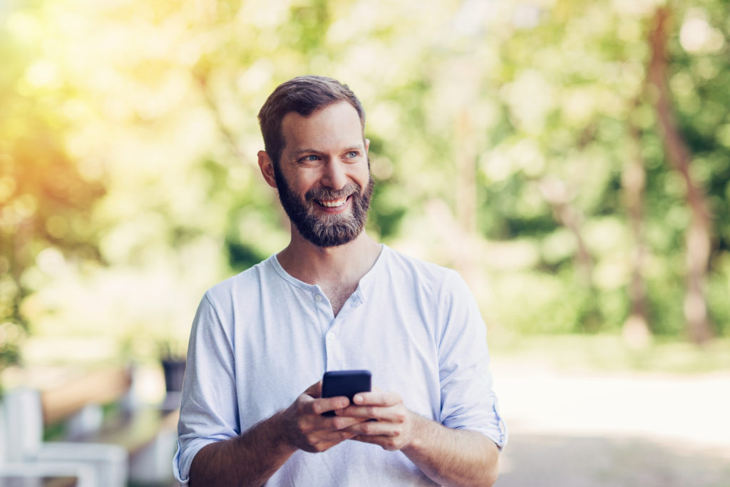 A bearded man in a white shirt smiles while holding a smartphone outdoors, perhaps checking GSMA IMEI services on his device. Sunlight filters through green trees in the background, creating a warm, blurred atmosphere. The man looks slightly to his left, appearing happy and relaxed.